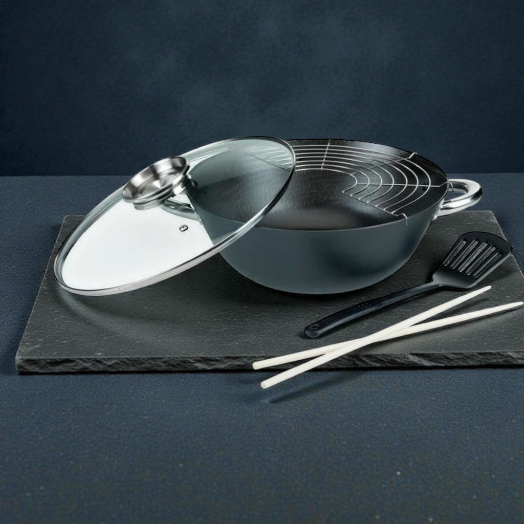 Schwarzer Wok mit Glasdeckel, Abtropfgitter und Küchenzubehör auf dunklem Untergrund