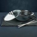 Schwarzer Wok mit Glasdeckel, Abtropfgitter und Küchenzubehör auf dunklem Untergrund